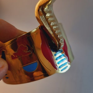 May include: A gold-colored bracelet with red, blue, and white enamel accents. The bracelet features a stylized lion design and a geometric pattern.