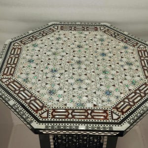 May include: An octagonal table with a decorative inlaid top. The surface features a mosaic pattern of white, brown, and iridescent pieces. The border is detailed with geometric designs. The table has a dark, possibly black, base.