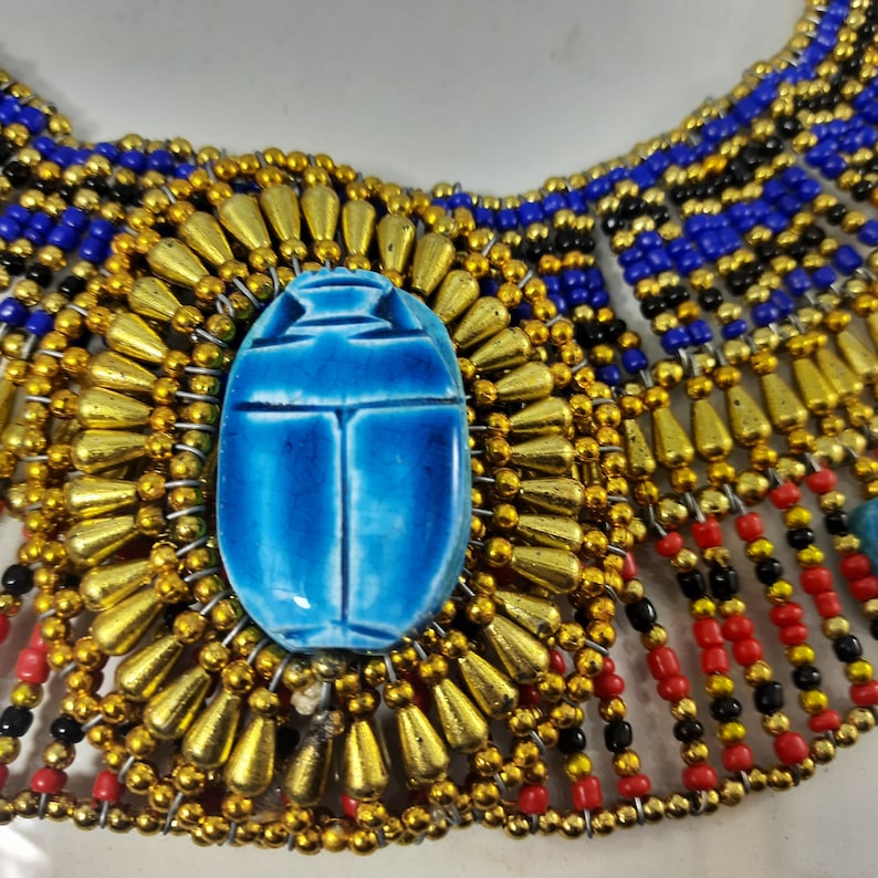 May include: A blue scarab beetle charm surrounded by gold beads, part of a beaded necklace with black, red, and blue beads.