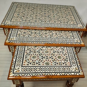 May include: Set of three nesting tables with intricate mosaic inlaid tops. The tables have a warm brown wooden frame and legs. The mosaic design features geometric patterns in shades of white, beige, brown, and blue. The tables are of varying sizes.