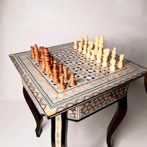 May include: A wooden chessboard table with intricate inlaid designs in shades of brown, white, and blue. The chessboard is set up with wooden chess pieces, some in the starting positions.
