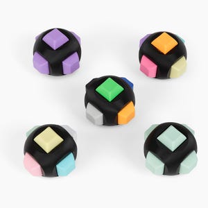 May include: A collection of five black, spherical puzzle toys. Each toy features colorful, geometric shapes in various colors, including purple, orange, pink, green, and blue. The toys are arranged on a white background.