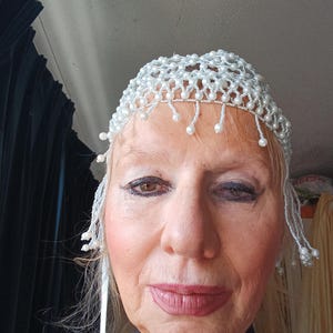 May include: A white beaded head covering with dangling pearl accents. The headpiece is made of small, round, white beads, and is designed to sit atop the head. The dangling accents add a touch of elegance.