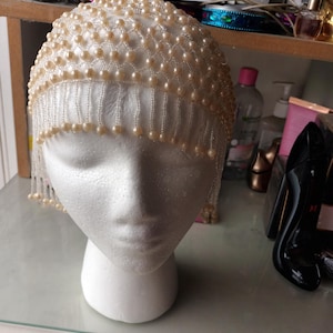 May include: A cream-coloured head covering, embellished with many small, round, faux pearls. The pearls are arranged in a net-like pattern across the top and cascade down in fringe-like strands. The head covering is displayed on a white foam head.