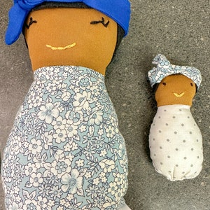 May include: Two handmade fabric dolls. The larger doll has brown skin, black hair, a blue headband, and a floral dress. The smaller doll has brown skin, a floral headband, and a white dress with gray polka dots. Both dolls have embroidered facial features.