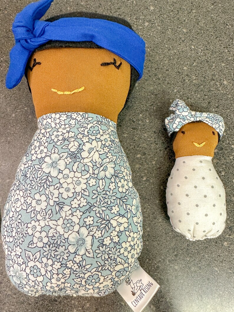 May include: Two handmade fabric dolls. The larger doll has brown skin, black hair, a blue headband, and a floral dress. The smaller doll has brown skin, a floral headband, and a white dress with gray polka dots. Both dolls have embroidered facial features.