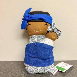 May include: A handmade fabric doll depicting a mother and child. The mother doll has brown skin, a blue headband, and a floral patterned dress. The child doll is held in a blue wrap. The dolls have embroidered facial features.