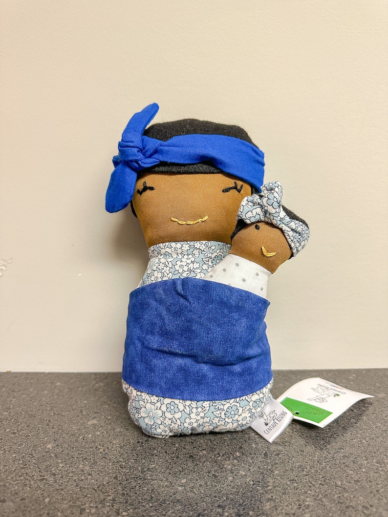 May include: A handmade fabric doll depicting a mother and child. The mother doll has brown skin, a blue headband, and a floral patterned dress. The child doll is held in a blue wrap. The dolls have embroidered facial features.