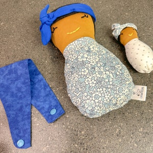 May include: Three handmade fabric dolls with brown skin and embroidered smiles. One doll wears a blue headband and a floral print dress. A smaller doll is wrapped in a white fabric with gray polka dots. A blue fabric strap with two buttons is also present.