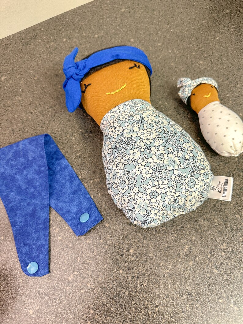 May include: Three handmade fabric dolls with brown skin and embroidered smiles. One doll wears a blue headband and a floral print dress. A smaller doll is wrapped in a white fabric with gray polka dots. A blue fabric strap with two buttons is also present.
