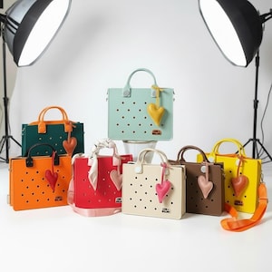 Könnte beinhalten: Eine Sammlung farbenfroher Handtaschen in verschiedenen Farbtönen, darunter Orange, Grün, Rot, Beige, Braun, Gelb und Türkis. Jede Tasche hat einen Griff, einen abnehmbaren Riemen und einen herzförmigen Anhänger. Die Taschen haben ein perforiertes Design und den Text "LANE & WILLOW".