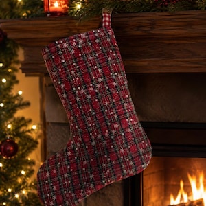 May include: A Christmas stocking featuring a red, green, and black plaid pattern with a white snowflake print throughout. The stocking includes a small red and white checkered fabric loop at the top for hanging, providing a festive holiday look.