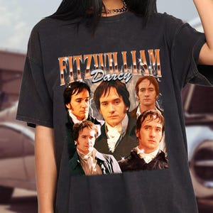 May include: Black t-shirt featuring a vintage-style graphic of Fitzwilliam Darcy from Pride and Prejudice. The design includes the name "Fitzwilliam Darcy" in orange and white lettering above a collage of portraits. The shirt is worn by a person.