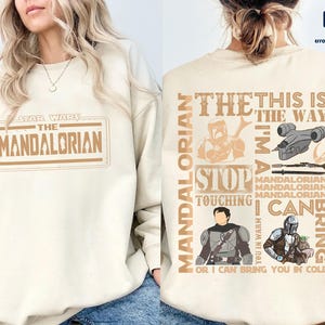 Puede incluir: Sudadera color crema con el logotipo y gr&aacute;ficos de "Star Wars: The Mandalorian". La parte delantera muestra el t&iacute;tulo del programa en un dise&ntilde;o rectangular. La parte trasera tiene ilustraciones de personajes y frases como "This is the Way".