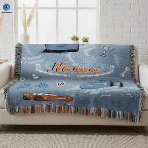 A blue and white throw blanket with a Cars-themed design, featuring a race car, tires, and other car-related elements. The blanket has a name printed on it, and it is placed on a white couch.