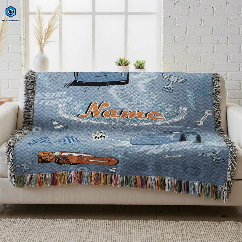 A blue and white throw blanket with a Cars-themed design, featuring a race car, tires, and other car-related elements. The blanket has a name printed on it, and it is placed on a white couch.