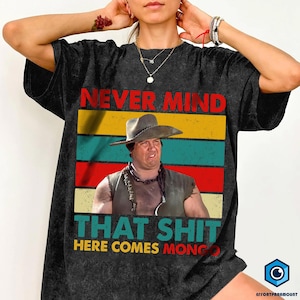 May include: Black t-shirt with a graphic featuring a man in a cowboy hat and the text "NEVER MIND THAT SHIT HERE COMES MONGO" in red, yellow, blue, and green stripes. The shirt has a vintage, worn look.