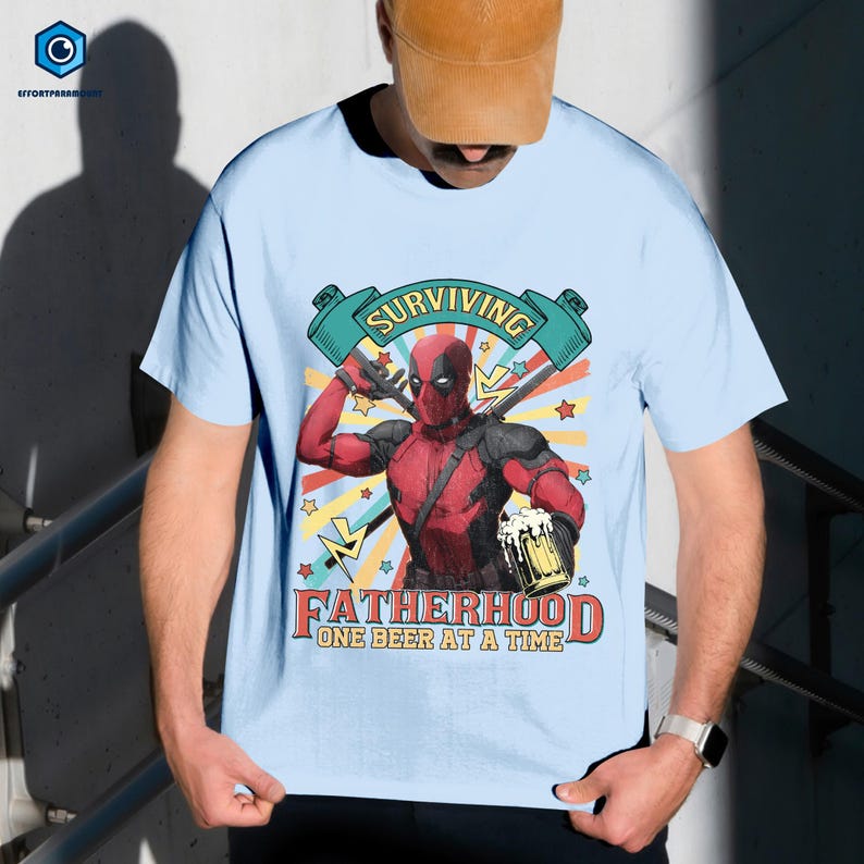 A person wearing a light blue t-shirt with a graphic design featuring a character from the Marvel Cinematic Universe, specifically the character Deadpool. The design includes the text "Surviving Fatherhood" and "One Beer at a Time", suggesting a humorous take on fatherhood.