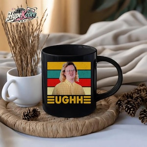 May include: Black ceramic mug featuring a retro design with a person wearing glasses and the word "EUGHH" in yellow. The design includes horizontal stripes in yellow, green, red, and blue. A white mug and decorative items are in the background.