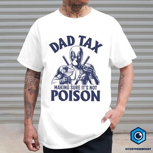 May include: White t-shirt featuring a graphic of a masked character holding a cocktail and a burger, with the text "DAD TAX MAKING SURE IT'S NOT POISON" in navy blue. The design is centered on the shirt.