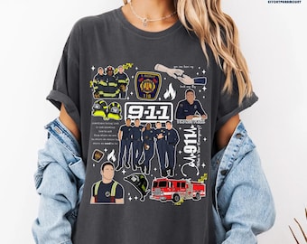 May include: Dark grey t-shirt with illustrations of firefighters, a fire engine, and the 9-1-1 logo. The design includes the text "What is your emergency?" and other related graphics.