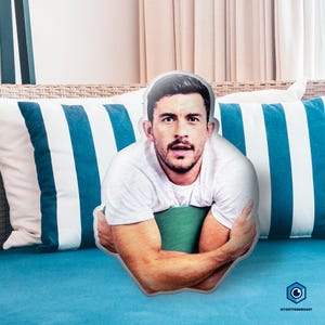 A man with a beard is sitting on a blue couch, with a pillow featuring a photo of him on it.