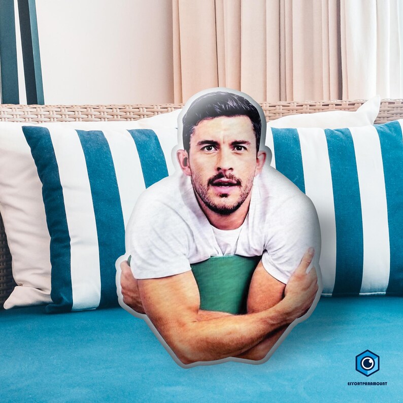 A man with a beard is sitting on a blue couch, with a pillow featuring a photo of him on it.