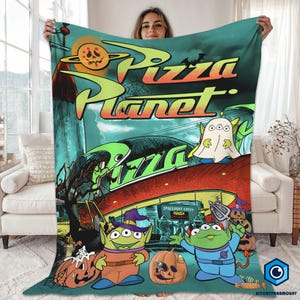 A woman is holding up a colorful blanket featuring an illustration of characters from the animated film "Pizza Planet".