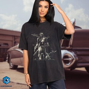 Pode incluir: Camiseta preta com um desenho gr&aacute;fico em preto e branco de um personagem Mandaloriano com uma lan&ccedil;a e um blaster. A camiseta tem mangas curtas e um corte folgado.