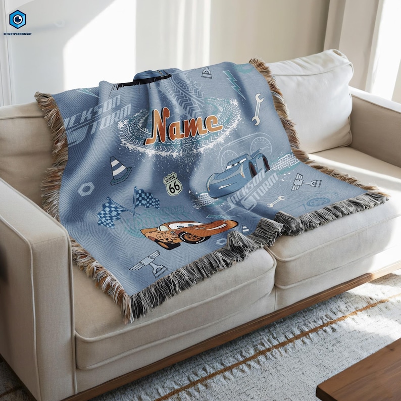 A blue blanket with a Cars-themed design, featuring various car-related elements such as a race car, a checkered flag, and a car with the number 66. The blanket is placed on a white couch, and the background appears to be a white wall.