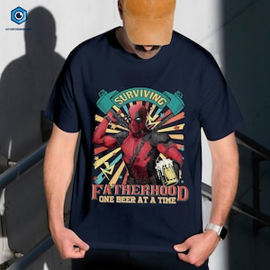 A person wearing a black t-shirt with a graphic design featuring a character from the Marvel Cinematic Universe, specifically the character Deadpool. The design includes the text "Surviving Fatherhood One Beer at a Time" and a banner with the character's face.