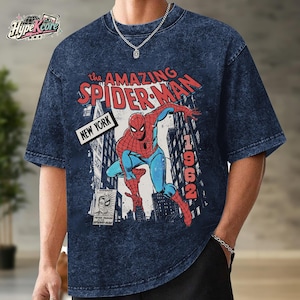 May include: A dark blue acid-wash t-shirt featuring a graphic of Spider-Man in a dynamic pose. The text reads The Amazing Spider-Man, New York, and 1962. A small inset image shows a split face of Peter Parker and Spider-Man with the text Peter Parker.