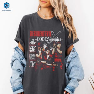 May include: Dark gray t-shirt featuring the text "RESIDENT EVIL - CODE: Veronica-" in red. The shirt has a graphic design with images of characters from the video game. The shirt is made of a soft material.