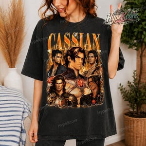 May include: A charcoal gray short-sleeve t-shirt featuring a large graphic print with the text CASSIAN in bold orange letters. The design displays five distinct anime-style illustrations of a male character with dark hair in various poses.