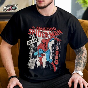 Może przedstawiać: Czarny t-shirt z kr&oacute;tkim rękawem z nadrukiem graficznym przedstawiającym Spider-Mana w czerwono-niebieskim kostiumie. Wz&oacute;r zawiera tekst "the AMAZING SPIDER-MAN", "NEW YORK" oraz "1962" na tle miejskich budynk&oacute;w i kadru z komiksu.
