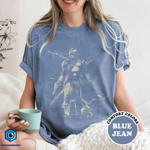 Pode incluir: Camiseta azul jeans com um gr&aacute;fico de um personagem com capacete e uma criatura pequena. A camisa tem mangas enroladas e as palavras "COMFORT COLORS" e "BLUE JEAN" em um design circular.