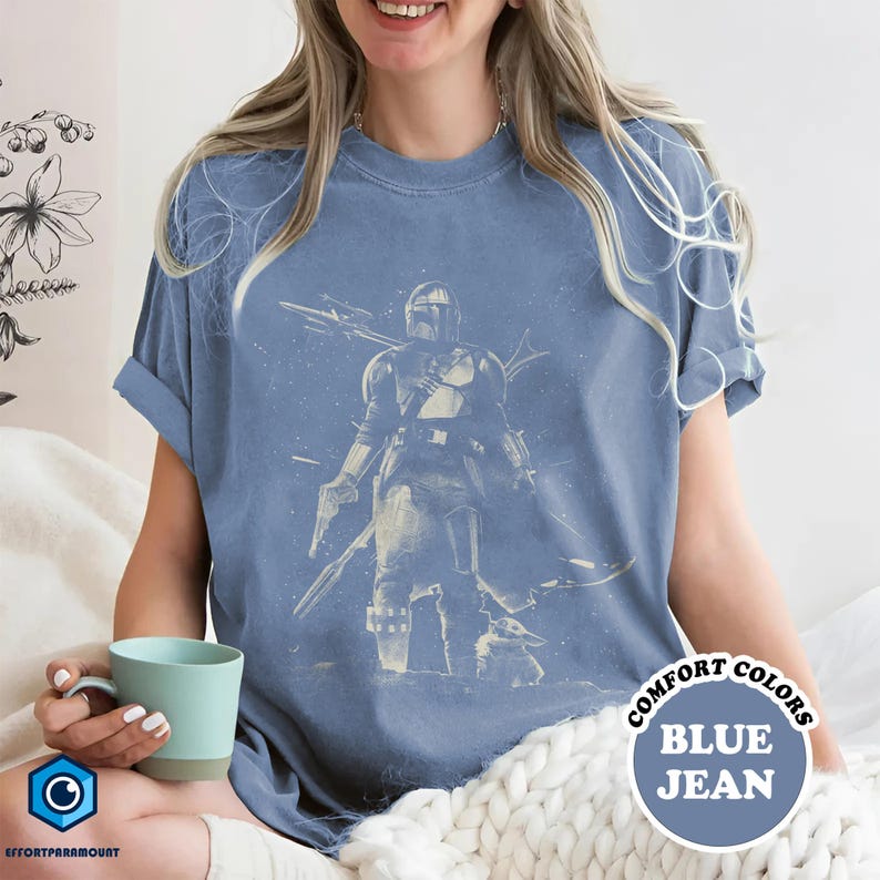 Pode incluir: Camiseta azul jeans com um gr&aacute;fico de um personagem com capacete e uma criatura pequena. A camisa tem mangas enroladas e as palavras "COMFORT COLORS" e "BLUE JEAN" em um design circular.
