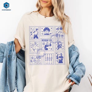 May include: Cream-colored t-shirt featuring a blue stamp-like design with images and text from the Resident Evil video game series. The design includes the text "RESIDENT EVIL" and "R.P.D."