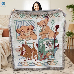 May include: Woven throw blanket featuring characters from The Lion King. The blanket has a cream background with images of Simba, Nala, Timon, Pumbaa, Rafiki, and Zazu. The border has a fringed edge with geometric patterns in shades of brown, green, and blue.