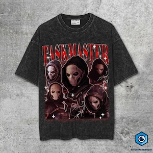 May include: Dark gray t-shirt featuring a graphic of masked figures with the word "TASKMASTER" in red. The design includes lightning bolts and stars, creating a vintage, distressed look. The shirt has a crew neck and short sleeves.