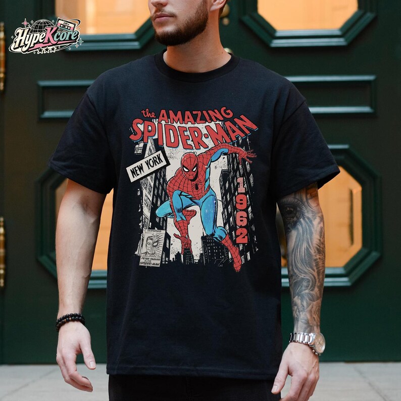 Może przedstawiać: Czarny t-shirt z kr&oacute;tkim rękawem z grafiką przedstawiającą Spider-Mana w dynamicznej pozie na tle panoramy Nowego Jorku. Napis głosi The Amazing Spider-Man, New York oraz 1962. Projekt zawiera mały wstawiony obraz przedstawiający twarz.