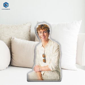 May include: A cut-out pillow featuring a smiling person in a light-colored linen shirt and pants, with sunglasses hanging from the shirt. The pillow is placed on a white couch with several pillows.