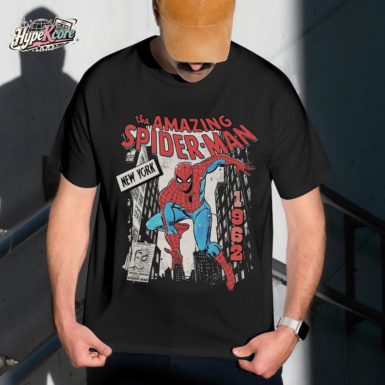 Może przedstawiać: Czarny t-shirt z kr&oacute;tkim rękawem z grafiką Spider-Mana w czerwono-niebieskim kostiumie. Tekst głosi The Amazing Spider-Man, New York oraz 1962. Projekt zawiera ilustrację w stylu komiksowym przedstawiającą budynki miejskie i mały plakat.