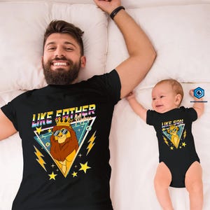 May include: Black t-shirt with a lion graphic wearing sunglasses and a crown, and the text "LIKE FATHER". A baby in a black onesie with a similar lion graphic and the text "LIKE SON". Both shirts have a retro design.