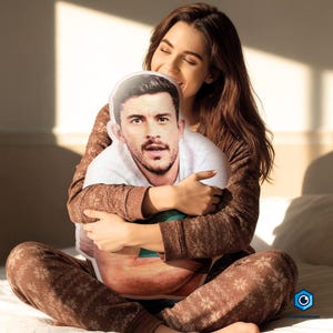 A woman is sitting on a bed, hugging a pillow with a man's face on it.