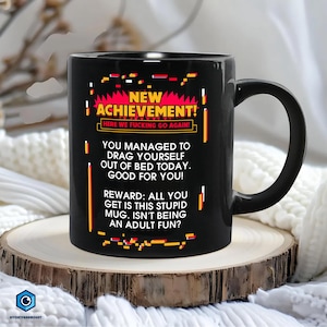 May include: Black ceramic mug with a humorous design. The mug features the text "NEW ACHIEVEMENT! HERE WE FUCKING GO AGAIN!" and other text. The mug is on a wooden surface.