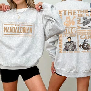 Puede incluir: Una sudadera gris claro con el texto "STAR WARS THE MANDALORIAN" en un dise&ntilde;o rectangular marr&oacute;n en la parte delantera. La parte trasera de la sudadera tiene un dise&ntilde;o con texto e im&aacute;genes relacionadas con la serie Mandalorian.