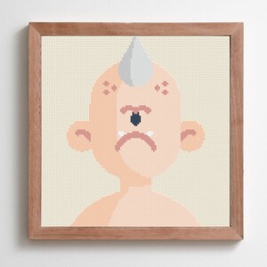 May include: A framed cross-stitch artwork featuring a stylized cyclops character. The character has peach skin, a single dark blue eye, and a gray horn. The artwork is set against a cream-colored background and framed in a light brown wood.