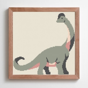 May include: A framed cross-stitch picture of a dinosaur. The dinosaur is mainly green with pink and black details, set against a cream background. The frame is made of light brown wood, adding a natural touch to the piece.