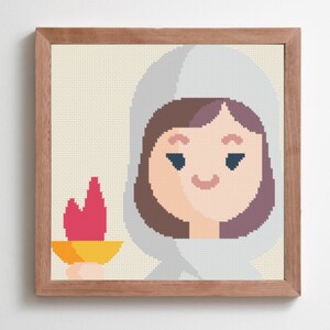 May include: Cross-stitch art of a person in a hooded cloak holding a torch. The person has brown hair and a light skin tone. The torch has a yellow base and a red flame. The artwork is framed in a light brown wooden frame.